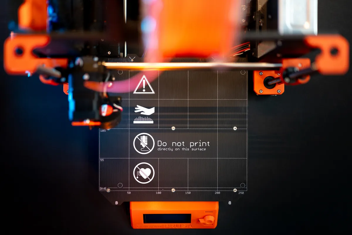 Draufsicht auf das beheizte Druckbett eines 3D-Druckers mit orangen Bauteilen und Warnsymbolen für heisse Oberfläche und Herzschrittmacher