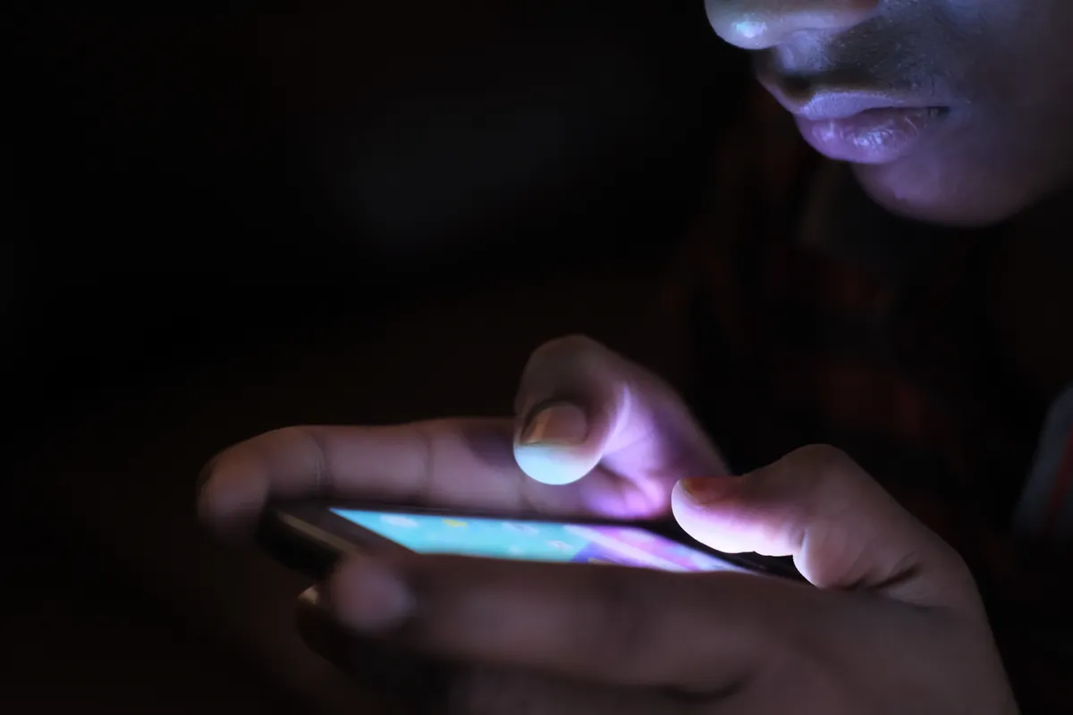 Zwei Hände tippen im Dunkeln auf einem leuchtenden Smartphone, das Display wirft violettes Licht auf Finger und Gesicht
